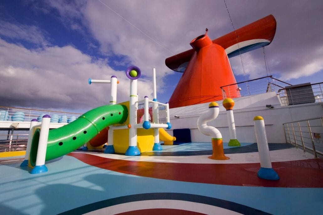 A colorful children's water play area on the Carnival Cruise Ship deck features various tubes, poles, and structures for climbing. The deck is red, white, and blue, with a large red funnel in the background under a partly cloudy sky.