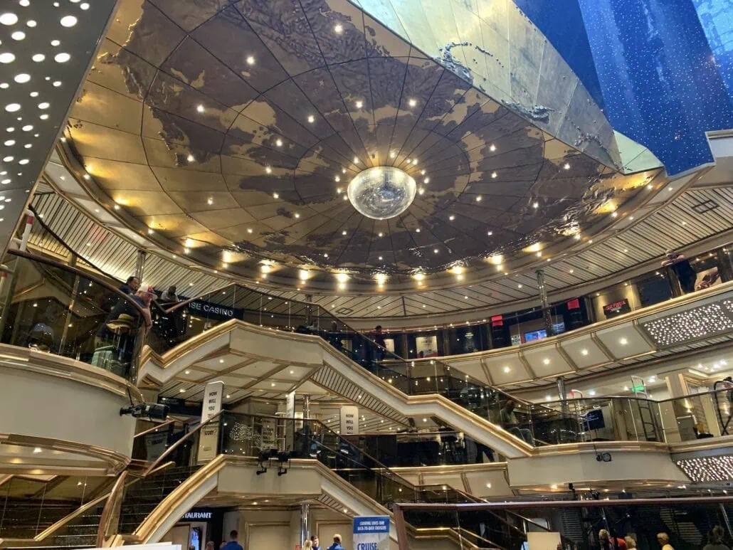 The interior of the multi-level shopping mall resembles the elegance of a grand Carnival Sunrise with its ornate gold ceiling featuring a world map design. The space is 2019 bustling, filled with people on escalators against a backdrop of vibrant retail signage and decorative lighting.