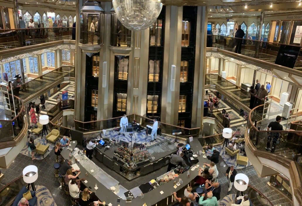 The 2019 Carnival Sunrise boasts a large, multi-level atrium where a central bar is surrounded by patrons at tables. A shiny disco ball glitters above, while ornate railings add elegance and shops entice in the background.