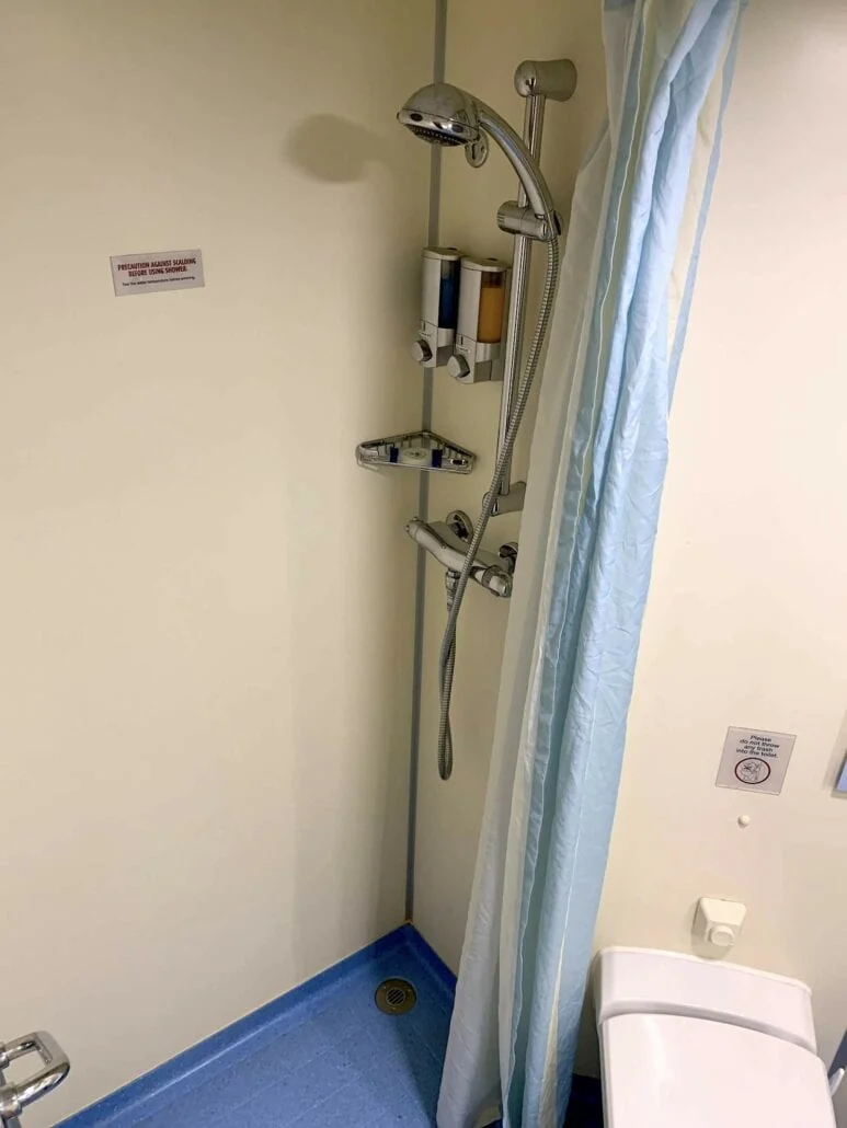 A small bathroom aboard the Carnival Sunrise features a shower area with a handheld showerhead, soap dispensers, and a blue-striped curtain. The blue floor complements the nautical theme perfectly, while a white toilet is partially visible to the right.
