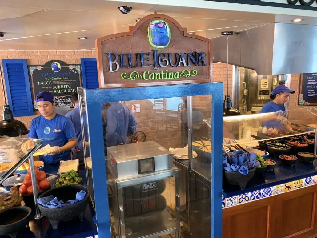 The image captures the Blue Iguana Cantina aboard the Carnival Sunrise. Two staff members are hard at work: one serves a customer while the other arranges toppings. Various ingredients, including tomatoes and sauces, are displayed beneath a decorative sign reading "Blue Iguana Cantina.