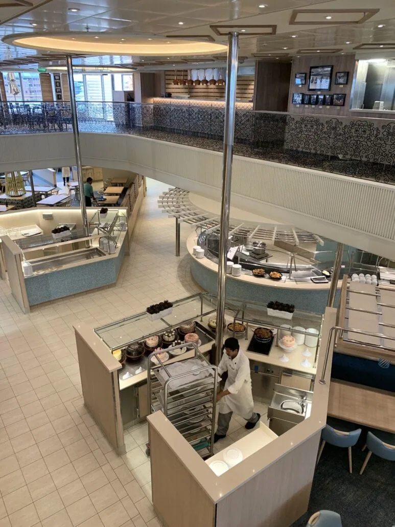 View of the multi-level cafeteria on the Carnival Sunrise, featuring various food stations. A staff member in a white uniform pushes a cart. Seating areas with tables and chairs are spread around, showcasing beige and white tones with decorative accents reminiscent of its 2019 design.