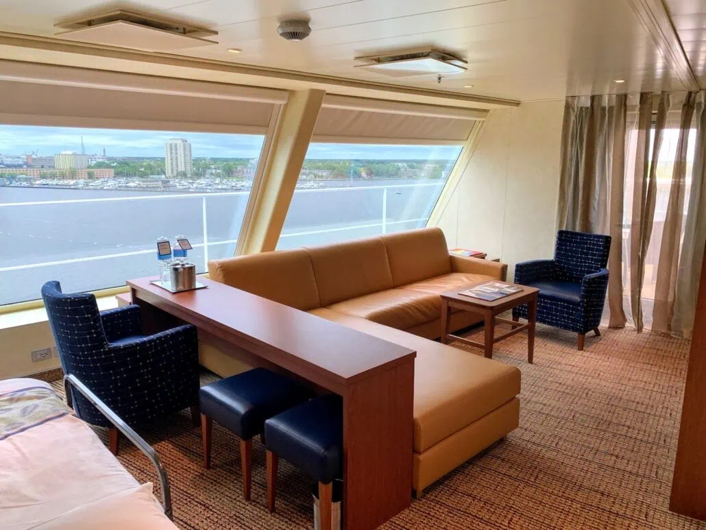 A spacious cabin on the 2019 Carnival Sunrise features a large window with city and water views. It includes a beige sectional sofa, two blue armchairs, a small rectangular table, and a wooden surface with two stools. Neutral tones and natural light create a cozy atmosphere, perfect for any ship review.