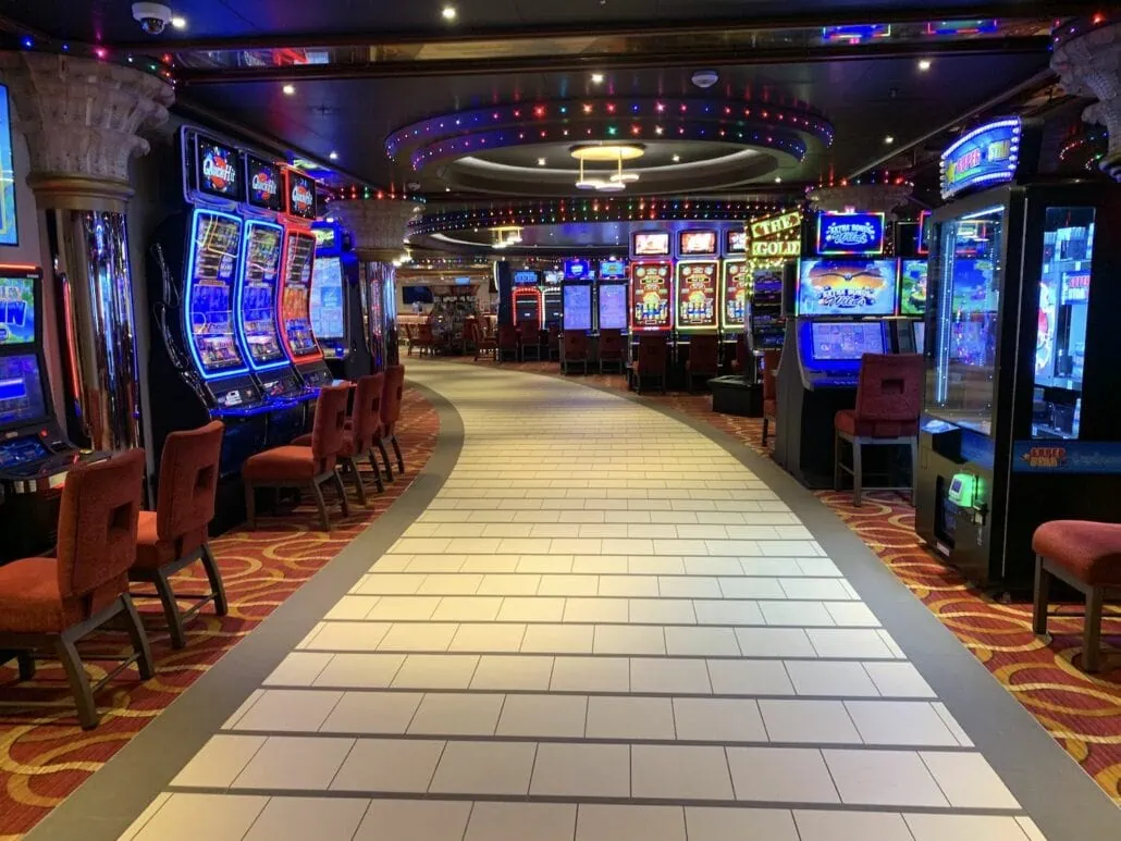 A brightly lit casino interior aboard the Carnival Sunrise features rows of slot machines lining a tiled walkway. Red upholstered chairs invite guests to try their luck, while circular lights and decorative pillars add an elegant touch.