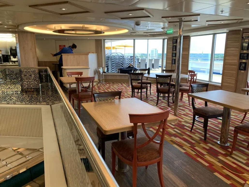 An empty dining area on the Carnival Sunrise cruise ship showcases wooden tables and chairs. Large windows offer an ocean view. A person in a blue shirt stands near a counter on the left, with stacked towels visible near the windows, capturing a serene snapshot of cruising elegance.