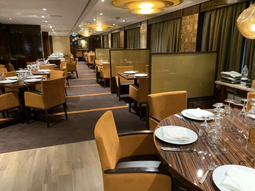 An elegant restaurant interior aboard the Carnival Sunrise features neatly arranged tables and orange upholstered chairs. Place settings include white plates and folded napkins. The room, reminiscent of 2019's classic designs, boasts warm lighting and a mix of wood and carpet flooring.