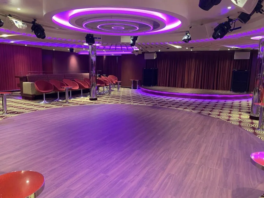 The modern dance floor of the 2019 Carnival Sunrise dazzles with purple lighting and circular patterns on a polished wooden surface. Red chairs, tables, and closed curtains form the background, while circular ceiling lights illuminate this vibrant space ready for an eclectic night at sea.