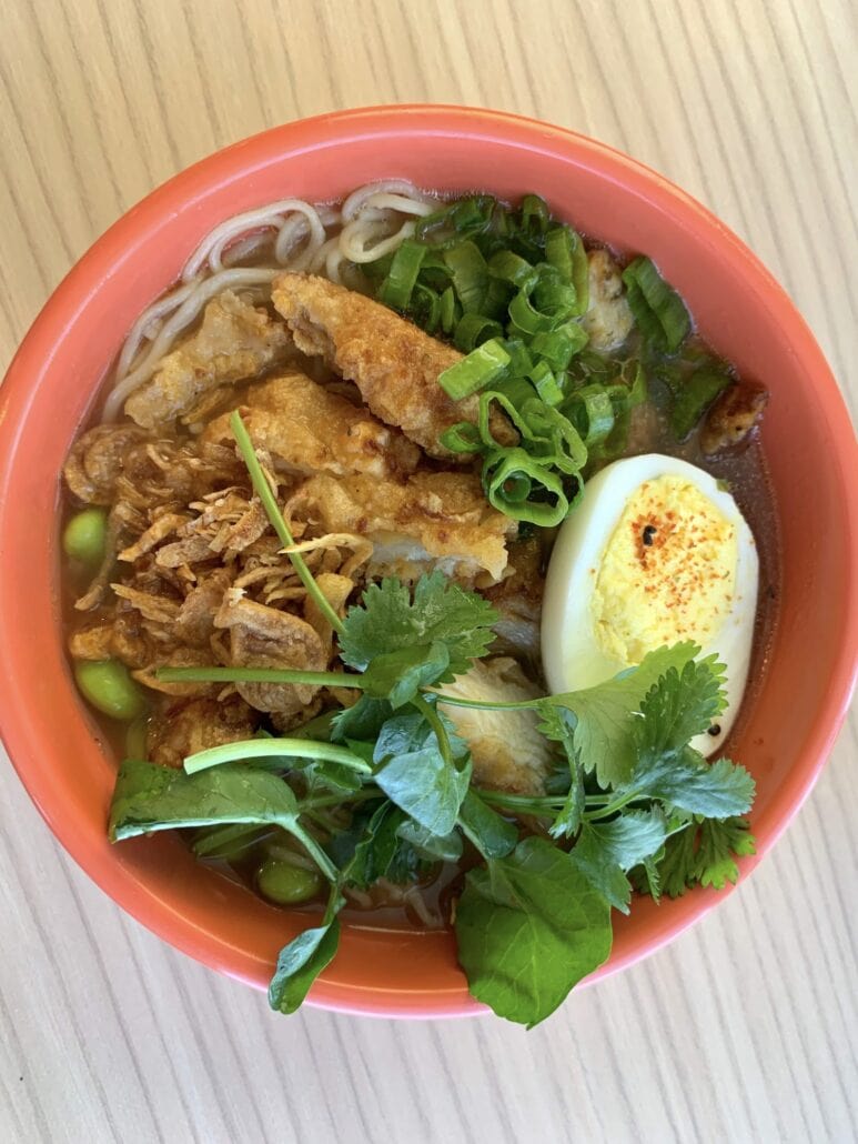 A vibrant bowl of noodle soup with fried tofu, sliced boiled egg, green onions, edamame, and fresh cilantro graces the table like a feast aboard the Carnival Sunrise in 2019. Served in an orange bowl on a wooden table, this dish is a flavorful voyage for your taste buds.