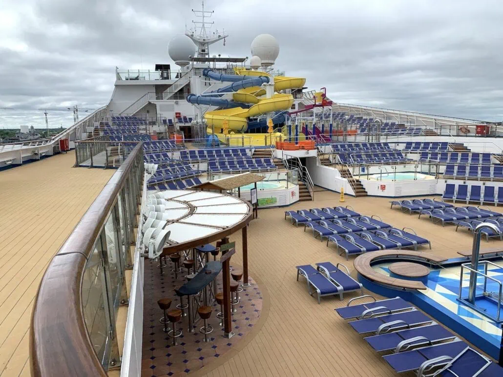 The image showcases the upper deck of the Carnival Sunrise cruise ship, featuring a yellow and blue waterslide, numerous empty lounge chairs, a circular bar with stools, and a small circular pool. The sky is overcast, adding to the serene ambiance of this 2019 voyage.