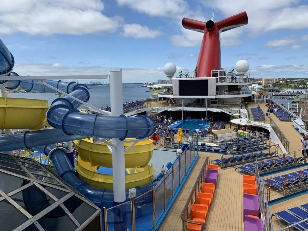 View of the Carnival Sunrise cruise ship deck, featuring a large waterslide, a red funnel, and colorful seating. The deck includes a pool area with loungers, a large screen, and multiple levels, set against the backdrop of the ocean and distant shoreline—truly an unforgettable 2019 experience.