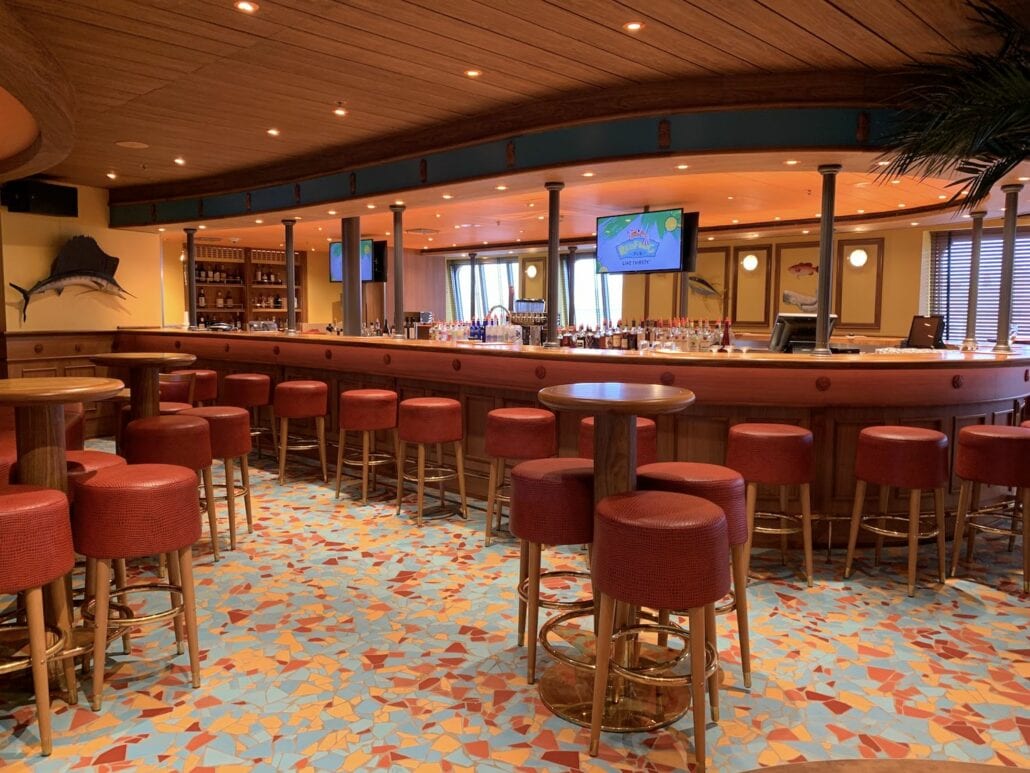 A bar with a nautical theme reminiscent of the Carnival Sunrise, featuring a long wooden counter surrounded by red cushioned stools. The floor boasts a colorful, mosaic-like pattern. Two TV screens are mounted above the bar, and shelves with bottles gleam in the background.