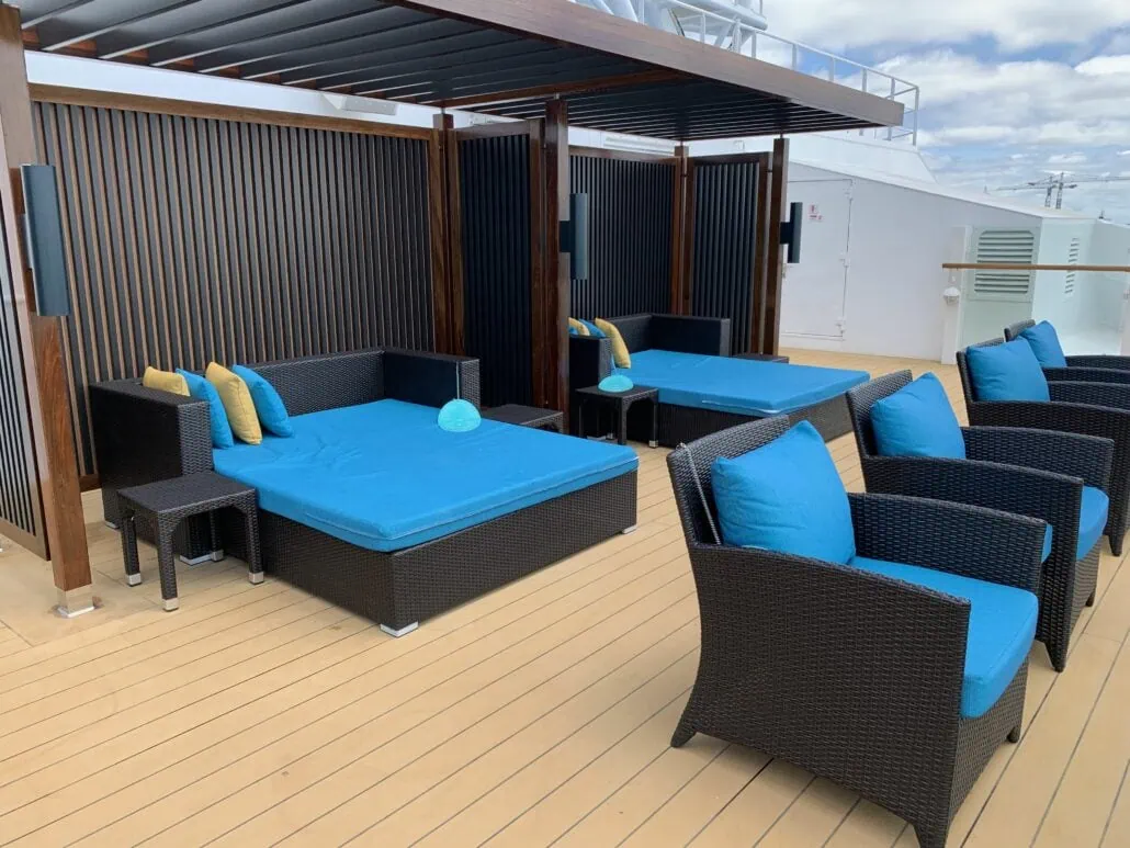 The outdoor deck area on the Carnival Sunrise features two dark wicker daybeds with blue cushions, side tables, and covered pergolas. Four blue-cushioned armchairs face the daybeds on a wooden plank floor, all under a partly cloudy 2019 sky.