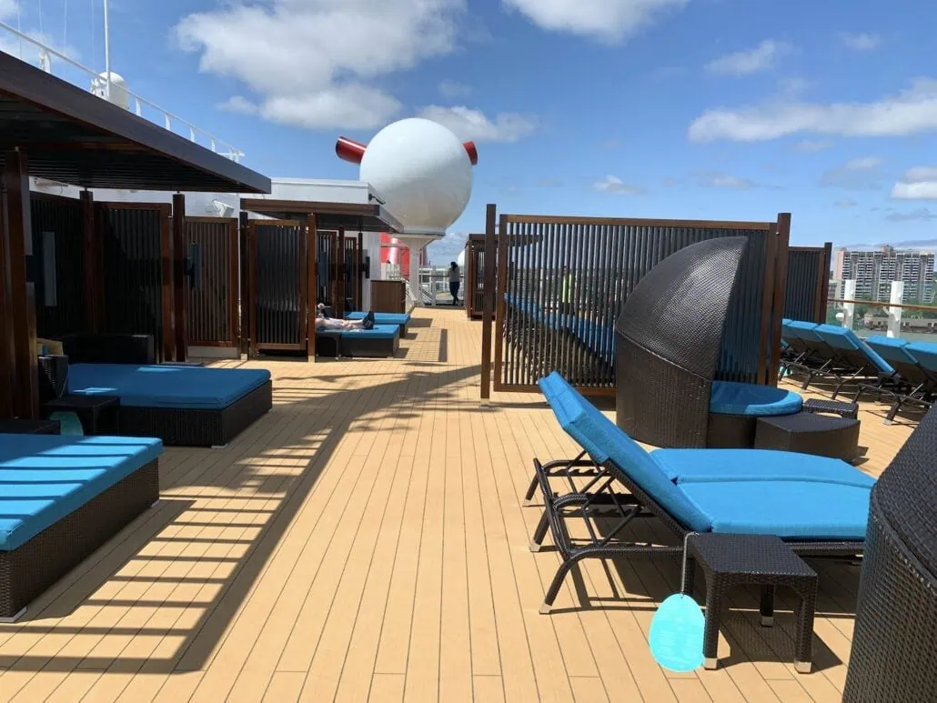 The sun deck on the Carnival Sunrise offers blue lounge chairs and dark wicker furniture on a light wood floor. In the background, enjoy a partial view of the ship's funnel and city buildings under a 2019 blue sky speckled with scattered clouds.