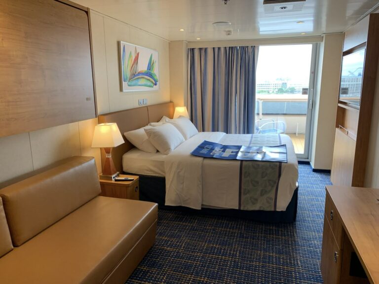 A well-lit cabin on Carnival Sunrise features a double bed with white linens and a blue runner, a beige sofa, a small wooden desk, and a balcony with sliding glass doors. A vibrant abstract painting hangs above the bed, contributing to the ship's welcoming atmosphere in this glowing 2019 debut.
