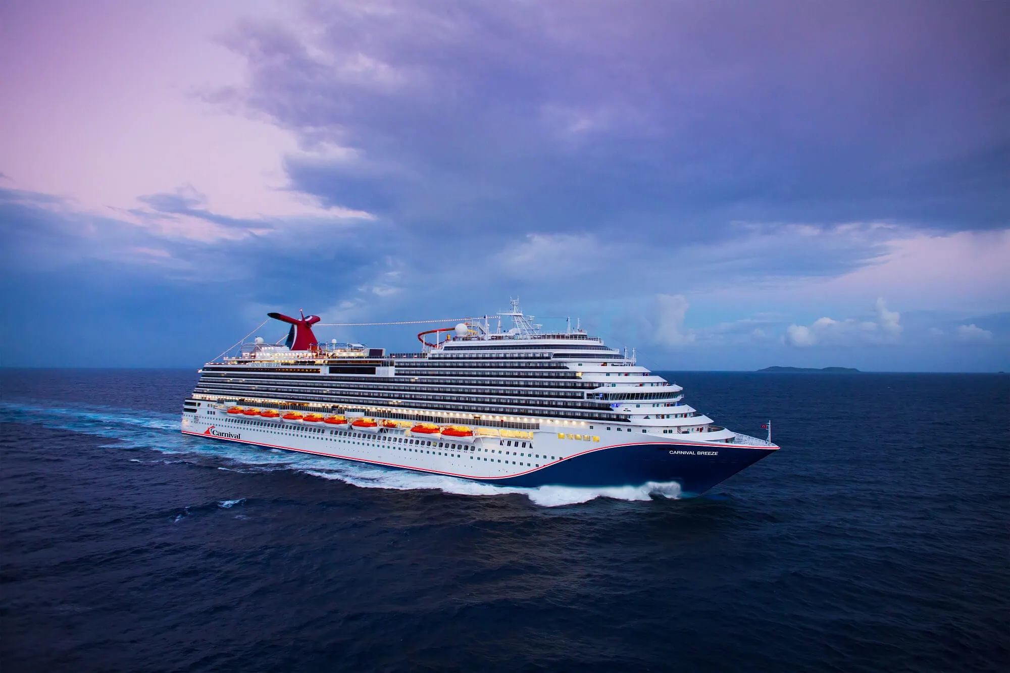 A Carnival Cruise ship sails across the open ocean under a cloudy sky. The brightly lit vessel showcases multiple decks, lifeboats lining its sides. As it glides through calm waters, the cruise leaves a gentle wake behind it, epitomizing a serene journey on the sea.