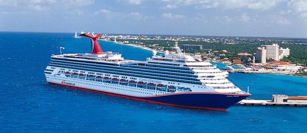 A majestic Carnival Cruise ship is docked at a port in the vibrant blue sea under a partly cloudy sky. The shoreline offers a picturesque town with mid-rise buildings and greenery, inviting exploration guided by the charm of seaside living.