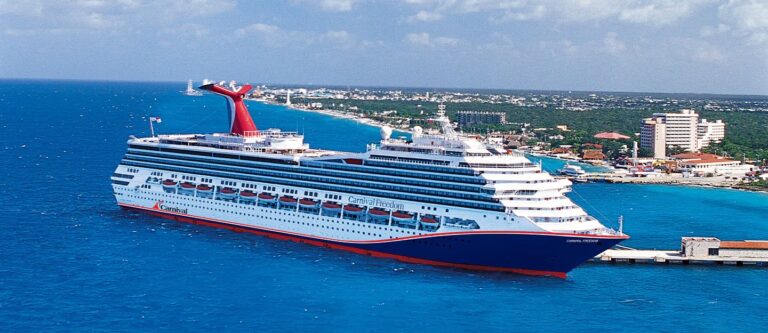A majestic Carnival Cruise ship is docked at a port in the vibrant blue sea under a partly cloudy sky. The shoreline offers a picturesque town with mid-rise buildings and greenery, inviting exploration guided by the charm of seaside living.
