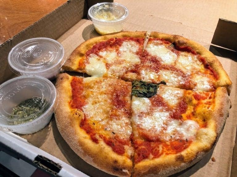 A partially sliced pizza with melted cheese and red tomato sauce on a thick crust is placed in a cardboard box. Next to it are three small plastic containers, each with different toppings or sauces, offering diverse dining options akin to the culinary delights aboard the Carnival Sunrise.