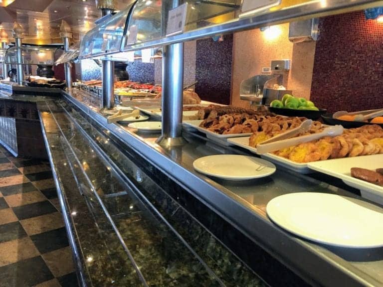 A buffet setup featuring a variety of foods. The foreground shows trays of pastries, fruit, and other dishes, with a generous spread of butter alongside. The background highlights more food trays under a glass guard along a long counter. The area is well-lit with a checkered floor, reminiscent of a cruise ship dining hall.
