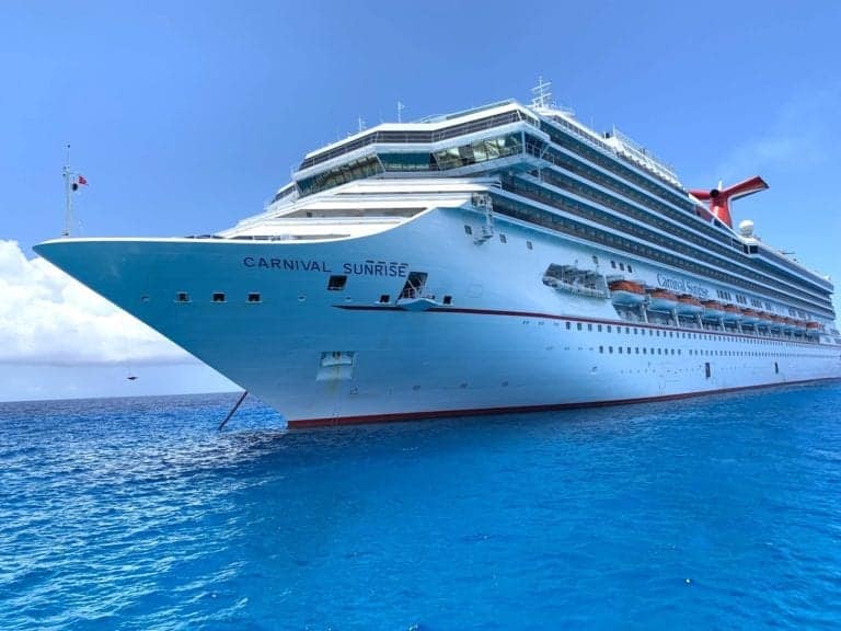 A large white cruise ship named "Carnival Sunrise," reviewed favorably in 2019, sails on a calm, bright blue ocean under a clear sky. The ship features multiple decks, windows, and a distinctive red and blue funnel at the rear.