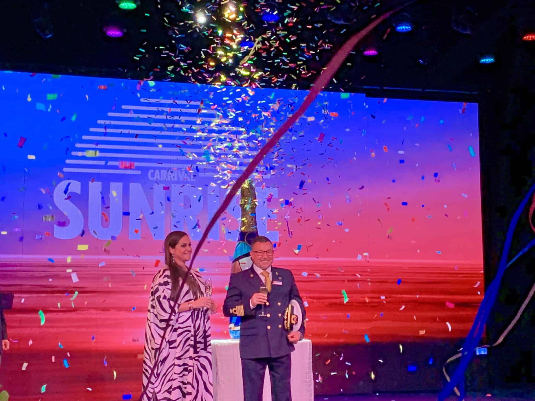 Two individuals are standing on a stage under a colorful shower of confetti and streamers. The background features a large screen displaying "Carnival Sunrise." One of them is in uniform, and they hold beverages together, smiling amidst the festive atmosphere reminiscent of New York City's grand celebrations.