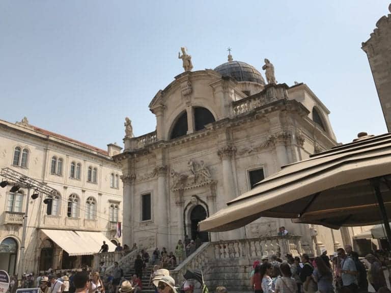 A large, historical stone building with a central dome and statues on the roof stands in the background. A crowd of people, including some fervent Game of Thrones Finale fans, is gathered in front of the building, with some shaded by a structure on the right. A smaller building is visible on the left.