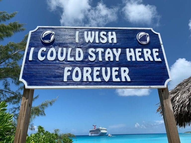A wooden sign with the text "I wish I could stay here forever" stands against a backdrop of the blue sky, ocean, and the Carnival Sunrise cruise ship near Half Moon Cay. The sign is adorned with two seashell icons on the top corners. In the foreground are green plants and a thatched structure.