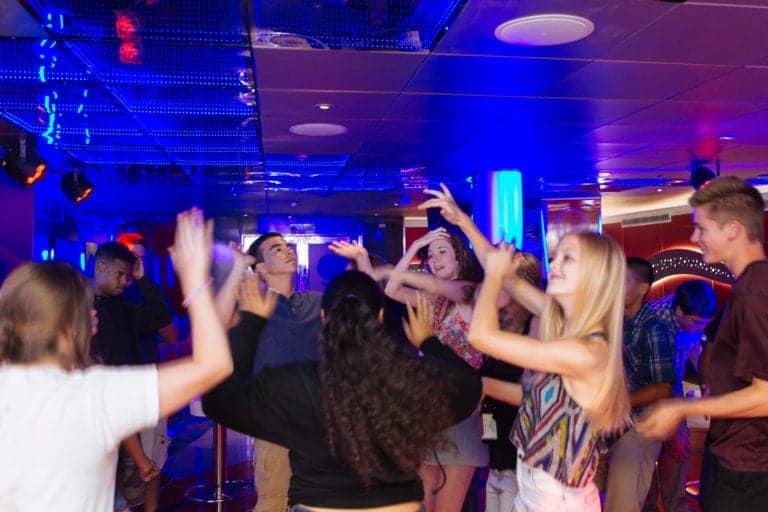 A group of people are dancing together in a club-like setting on a Carnival Cruise Line, with blue and purple lighting. They have their hands raised and seem to be enjoying themselves. The background features a ceiling with reflective panels and wall decorations.
