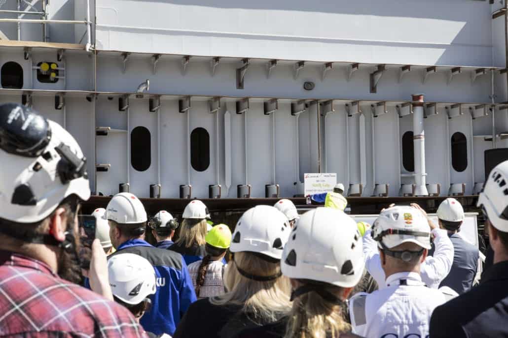 A group of people wearing helmets and safety gear are gathered and facing a large industrial structure. One person in a helmet is holding a sign, while others appear to be taking photos or observing. The background features metal beams and heavy machinery, evoking an industrial Mardi Gras atmosphere.