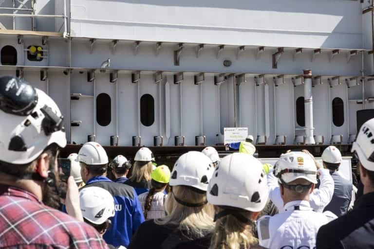 A group of people wearing helmets and safety gear are gathered and facing a large industrial structure. One person in a helmet is holding a sign, while others appear to be taking photos or observing. The background features metal beams and heavy machinery, evoking an industrial Mardi Gras atmosphere.