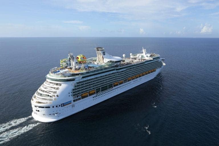 A large cruise ship sails on a calm sea under a partly cloudy sky. The ship has multiple decks with balconies and various recreational areas, including water slides and a colorful sphere. Despite its amenities, the memory of a toddler's fatal cruise ship fall lingers somberly among the waves rippling around the vessel.