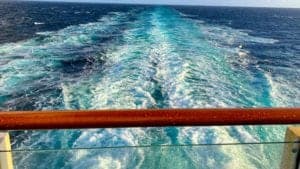 View from the back of a world cruise ship showing a frothy and turbulent wake trailing into the deep blue ocean, creating white and turquoise patterns. A wooden railing is visible in the foreground—a mesmerizing sight on this 120-night bucket list journey.