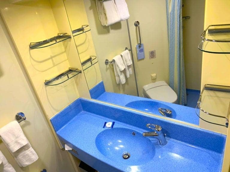 A small bathroom with a blue countertop and sink, a mirror above the sink, and yellow walls resembling those on a Carnival Cruise Line. Towels hang on racks beside and above the sink. The toilet is visible in the background next to a shower area with a curtain.