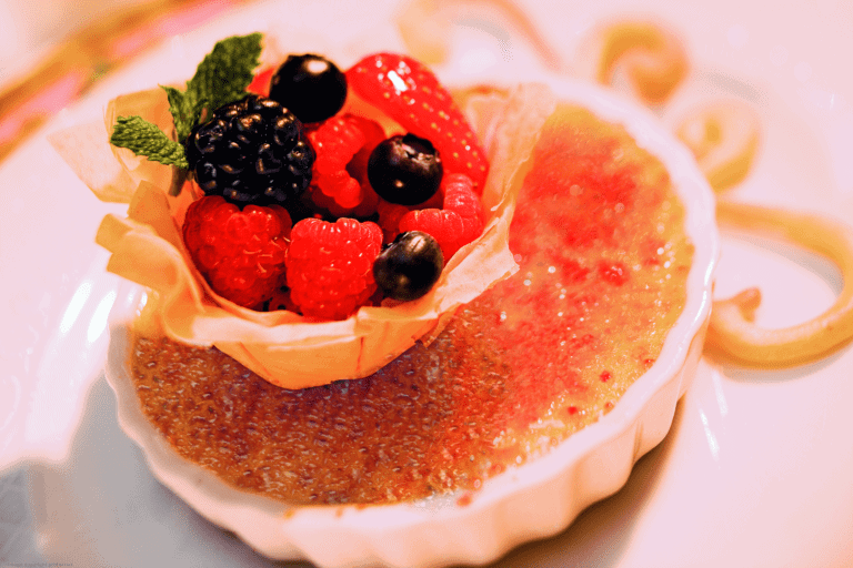 A dessert plate from Norwegian's Le Bistro MENU features a crème brûlée topped with a pastry cup filled with assorted berries, including raspberries, strawberries, blueberries, and blackberries, all garnished with a mint leaf and decorative drizzle around the dessert. Enjoy this specialty restaurant delight.