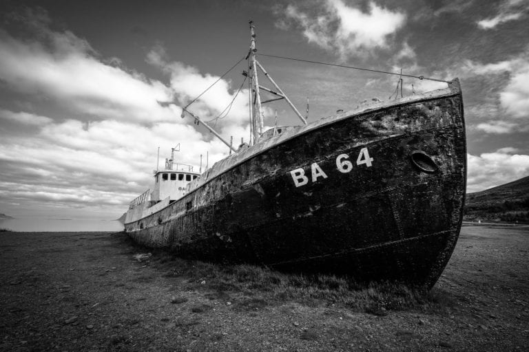 A black and white photo of an old, rusted ship beached on a rocky shore under a cloudy sky. The ship, once part of the brief history of gambling ships, has "BA 64" painted on its side. Vegetation is sparse, and the landscape appears desolate.