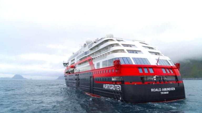 The pristine cruise ship "Roald Amundsen," proudly christened and bearing the "Hurtigruten Tromsø" on its stern, sails through icy waters. With white and red accents, it navigates under cloudy skies with distant landmasses coming into view, echoing an expedition to Antarctica.