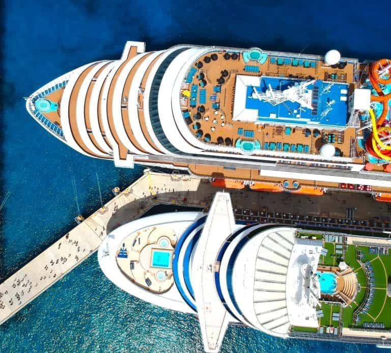 An aerial view of two docked Carnival Corporation cruise ships at a bustling harbor. The ships, side by side, showcase their decks with pools, lounging areas, and various facilities. The dock extends between them with several people visible on the pier, as the ocean investment surrounds the scene.