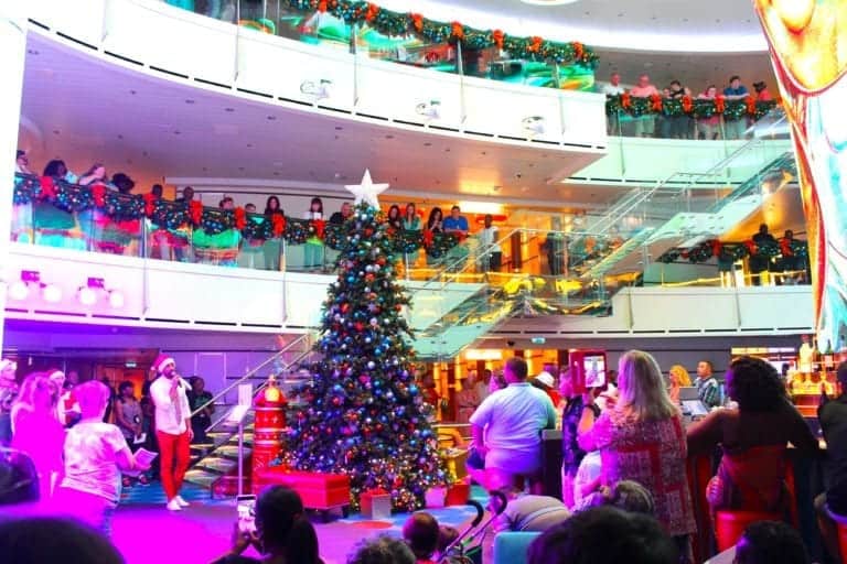 A multi-level indoor space is decorated for the 2019 Christmas carnival, with a large Christmas tree in the center. People are gathered around the tree and standing on balconies. Green garlands with red ribbons are hung on the railings. The area is brightly lit, reflecting an organized decorating schedule.