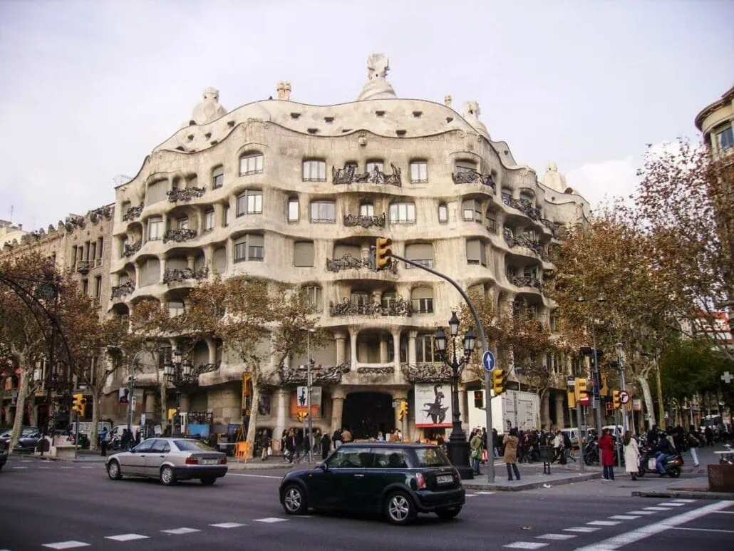 A street view of Casa Milà in Barcelona, Spain, showcasing its unique undulating stone facade and wrought iron balconies, with cars and pedestrians at an intersection in the foreground—one of the must-see things to do for cruisers exploring the city's architectural wonders.