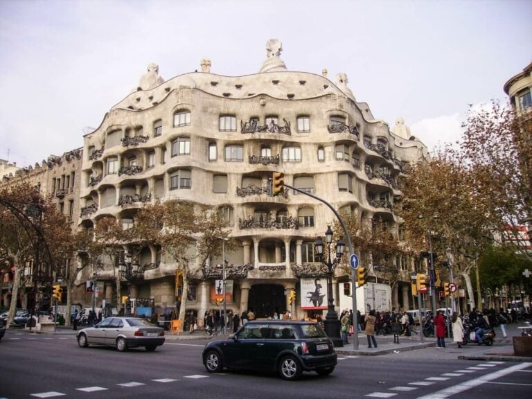 A street view of Casa Milà in Barcelona, Spain, showcasing its unique undulating stone facade and wrought iron balconies, with cars and pedestrians at an intersection in the foreground—one of the must-see things to do for cruisers exploring the city's architectural wonders.
