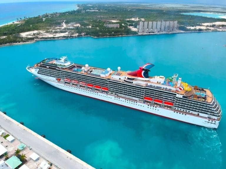 A large Carnival cruise ship is docked at a port in clear turquoise water. The ship has multiple decks and lifeboats along the sides. The shoreline visible in the background has buildings and greenery. The image is taken from a high vantage point.
