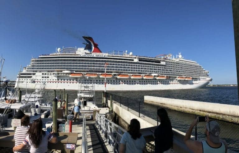 A large cruise ship named Carnival Breeze is docked at a port, reminiscent of the Rotterdam Report's detailed descriptions. Several people on a smaller dock in the foreground are watching and taking photos. The sky is clear and blue, with other smaller boats docked nearby, creating a picturesque farewell scene.