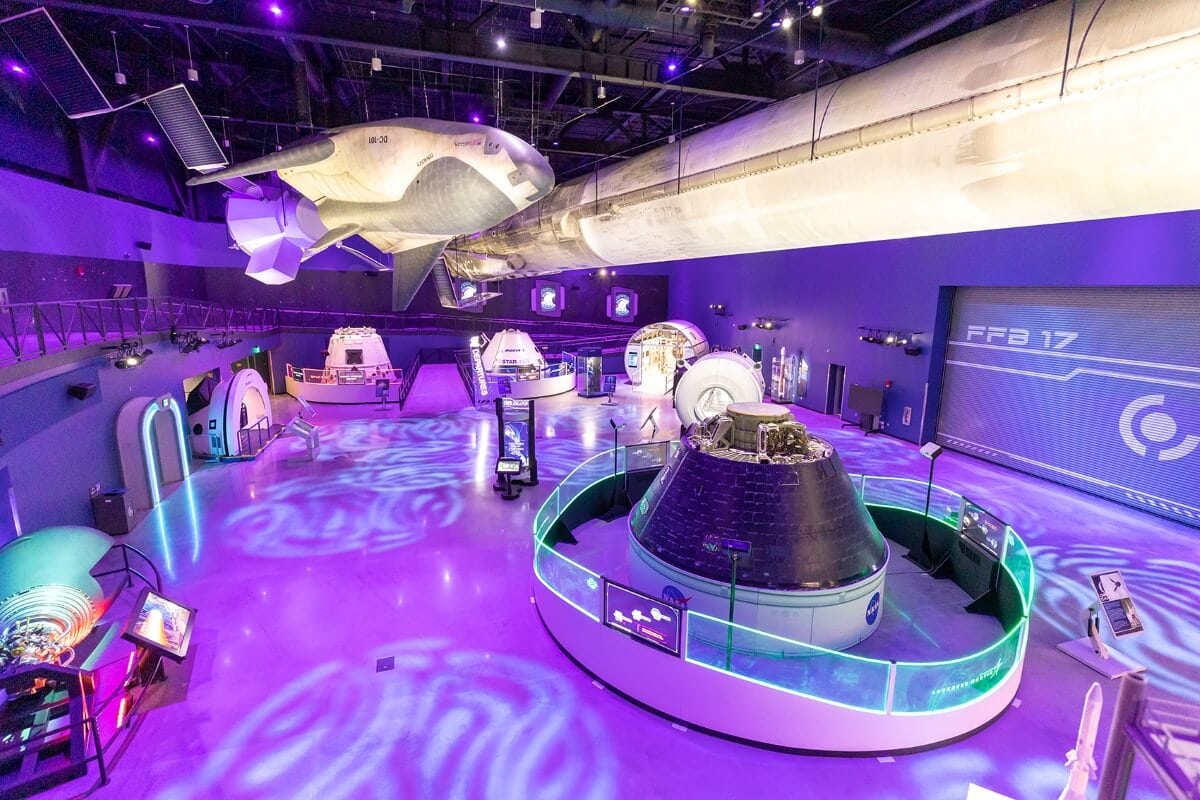 Interior of a space-themed museum exhibit featuring spacecraft displays, including a large model suspended from the ceiling, interactive stations, and an illuminated floor. For those cruising from Port Canaveral, this exhibit offers expert tips on navigating the cosmos.