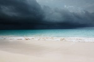 A sandy beach meets turquoise ocean waters under a dark, cloudy sky, suggesting an approaching storm. The waves gently crash onto the shore, reminiscent of the days past when Hurricane Dorian victims relied on relief amidst nature's fury.