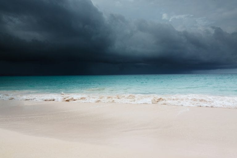 A sandy beach meets turquoise ocean waters under a dark, cloudy sky, suggesting an approaching storm. The waves gently crash onto the shore, reminiscent of the days past when Hurricane Dorian victims relied on relief amidst nature's fury.