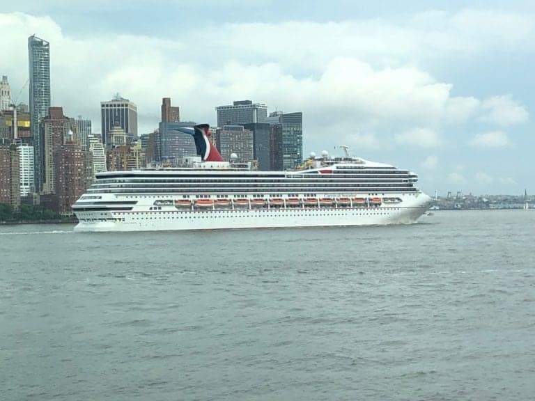 A large white cruise ship with red and blue accents is sailing on a body of water, with a city's skyline featuring tall buildings in the background. The sky is cloudy, setting the scene for discussions about major rule changes after reports of missing cruise workers.