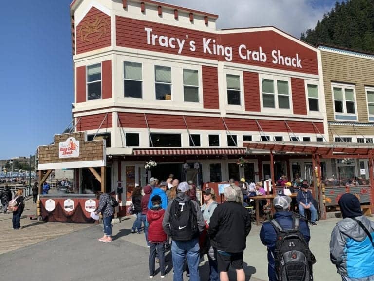 People stand in line outside Tracy's King Crab Shack, a red and white building with large windows. Located near the Juneau Cruise Port Guide, the busy sidewalk boasts signs of the restaurant's logo. The scene is framed by a mountainous landscape and a partly cloudy sky.