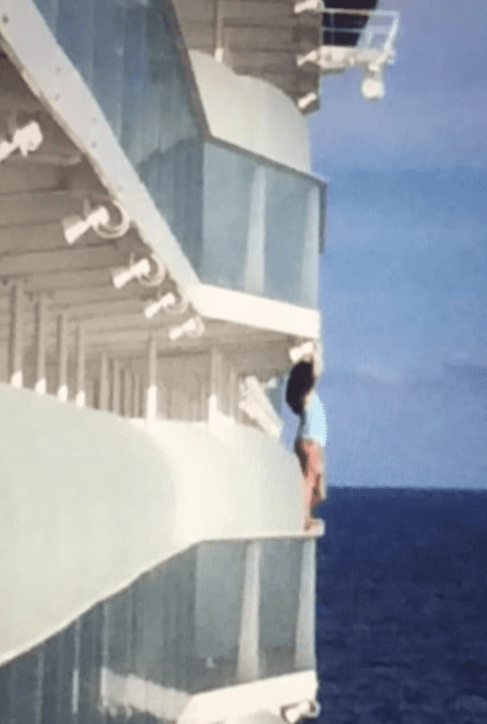 A passenger in a blue shirt and shorts is standing on the edge of a Royal Caribbean cruise ship balcony. The image shows multiple balconies and the ocean in the background. The person appears to be leaning outwards, facing away from the ship, possibly attempting a dangerous selfie.