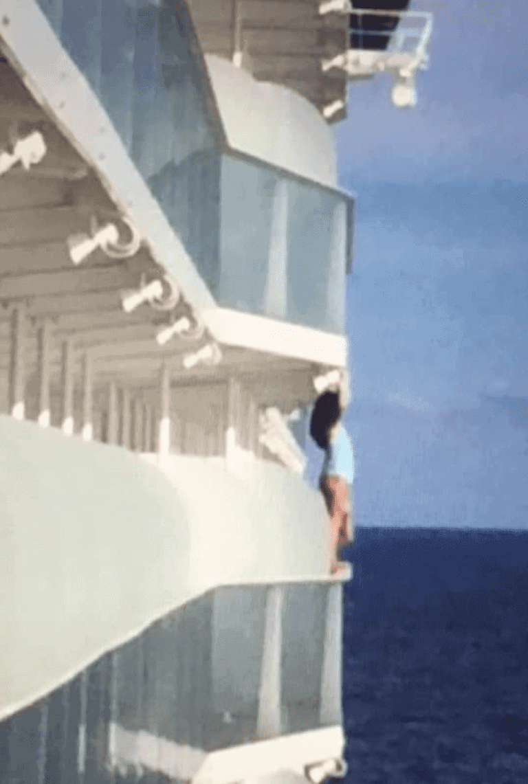 A passenger in a blue shirt and shorts is standing on the edge of a Royal Caribbean cruise ship balcony. The image shows multiple balconies and the ocean in the background. The person appears to be leaning outwards, facing away from the ship, possibly attempting a dangerous selfie.