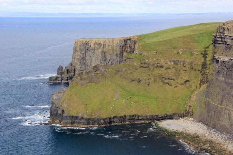 A view of a steep, rocky cliff covered in green grass along Dublin's coastline. The cliff faces the ocean, with waves crashing against its base. In the distance, the horizon meets the sky. The foreground features a rocky shoreline—one of Ireland's must-see sights and top things to do.